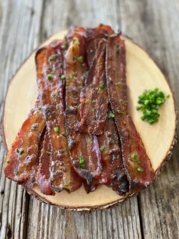 8 slices of cooked bacon with brown sugar, honey, and diced Jalapeno peppers. Bacon slices are on a round wood serving tray. There is a small pile of diced Jalapeno peppers on the tray. Tray is on a dark wood surface.