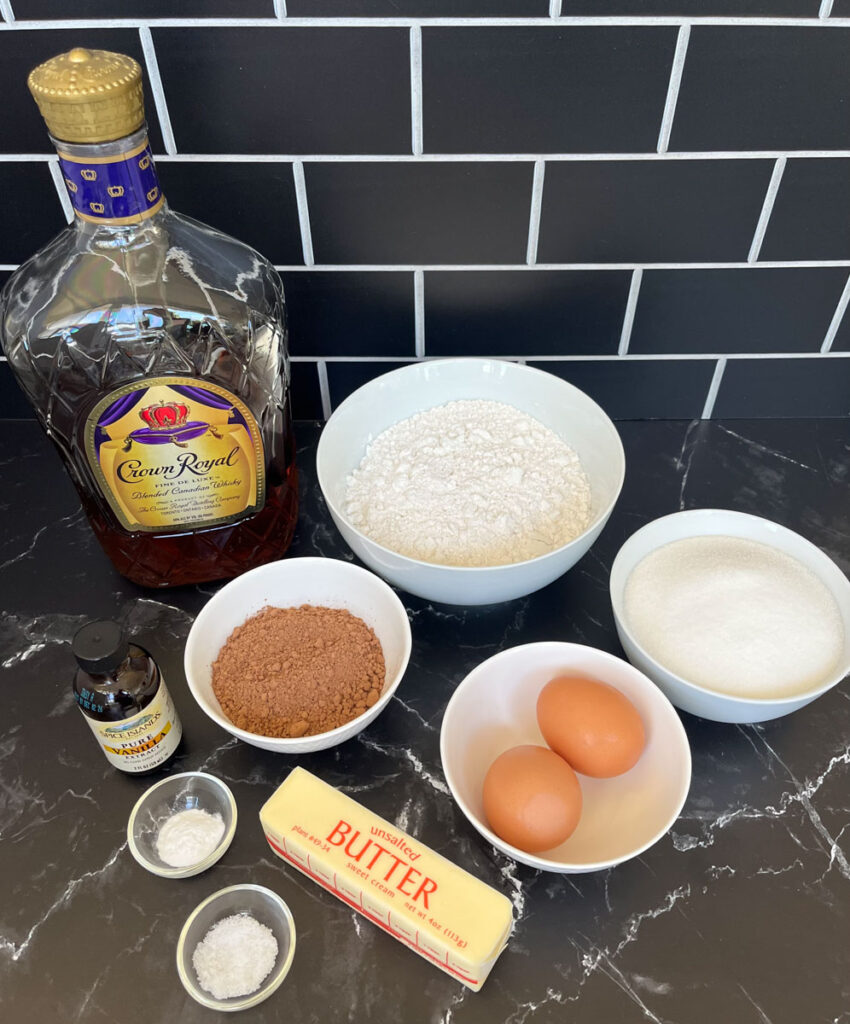 There is a bottle of Crown Royal. There is four white bowls. One bowl has flour in it, one with white sugar, one with coca powder, and one with two brown eggs in it. There is a stick of butter in front of the 4 bowls. There is a small glass bottle of pure vanilla extract and 2 small glass ramekins. One with baking powder and one with baking soda.