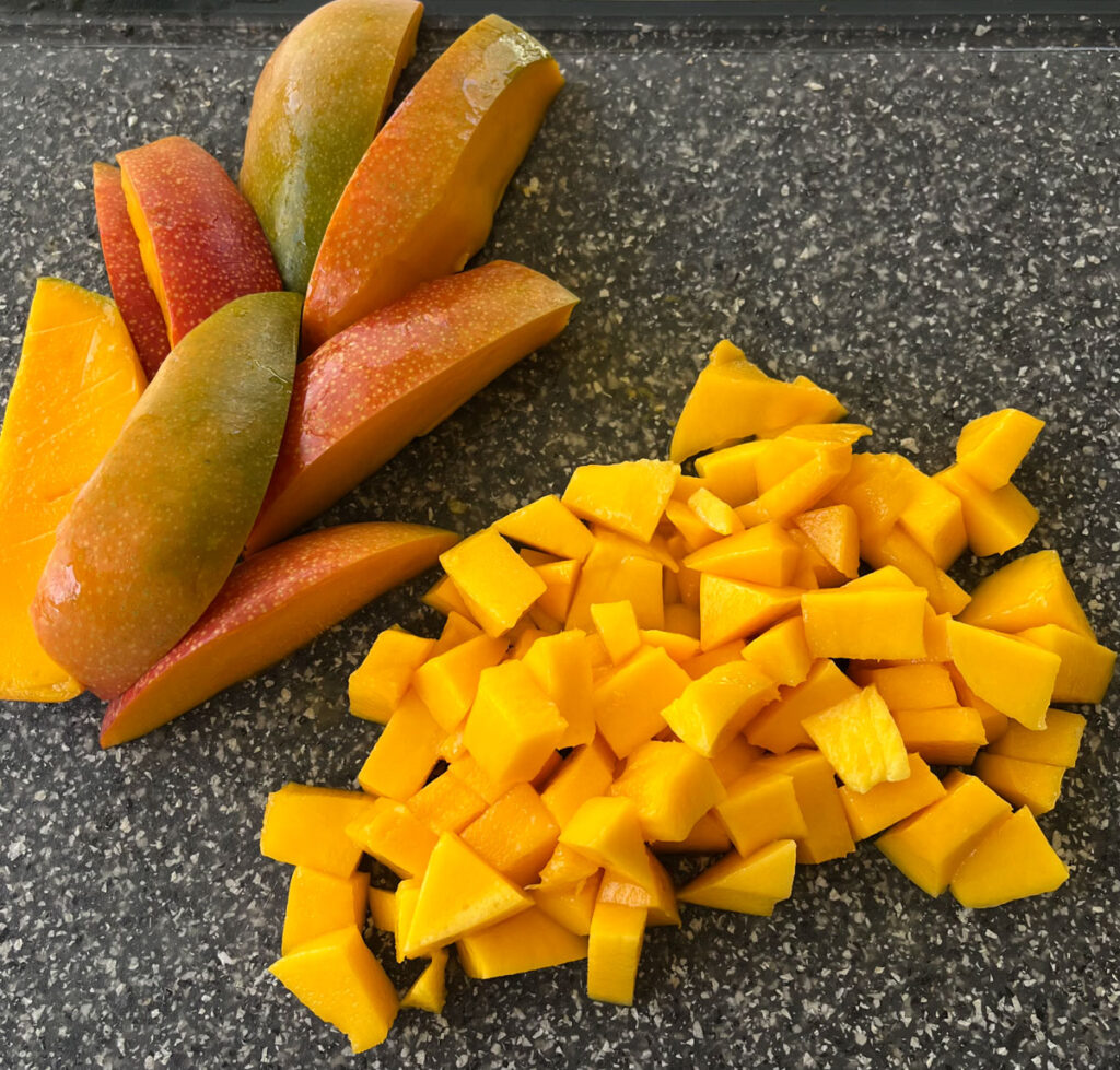 There are 8 slices of mango next to a pile of mango chunks on a gray and white speckled surface.