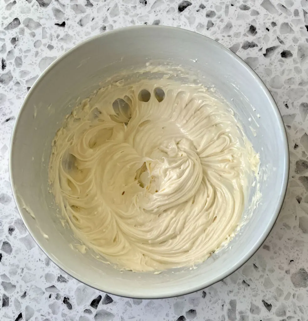 White mixing bowl with a cream cheese, vanilla, and sugar blended mixture. Bowl is on a white terrazzo surface.