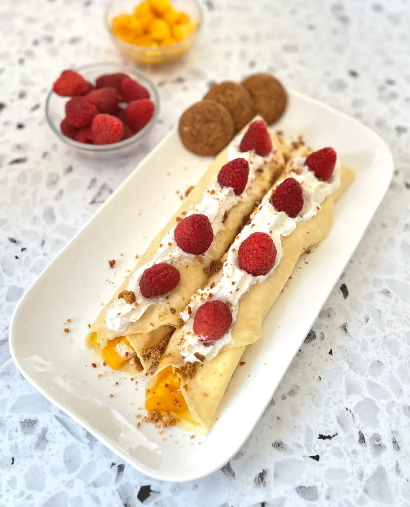 There are two rolled crepes with mango chunks in them. There are 4 raspberries on each crepe as well as whipped cream. The crepes are on a white rectangle plate. There are 3 small brown cookies on the plate. There is a small clear glass bowl of raspberries and another small clear glass bowl with cut up mango in it. Bowls are in the background. All items are on a white terrazzo surface.