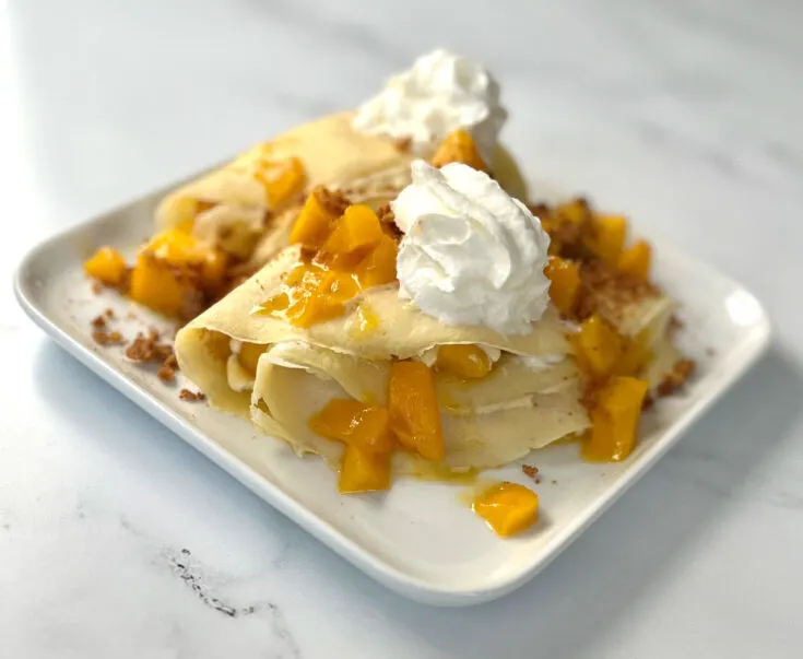 There are 2 triangle folded crepes with chunks of mango in them as well as on top of them. There is whipped cream and brown cookie crumbles on top of the crepes. Crepes are on a white square plate. Plate is on a white marbled surface.