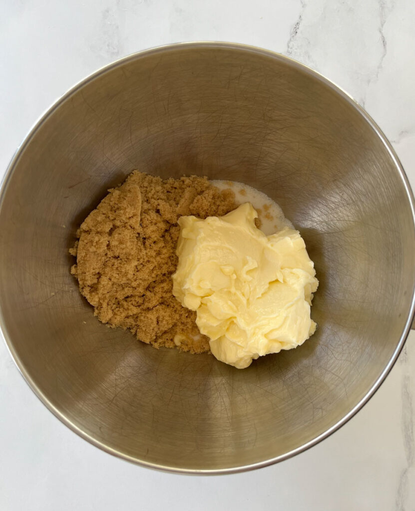Stainless steel mixing bowl with a pile of butter, light brown sugar, and white sugar in it. The bowl is on a white marble surface.