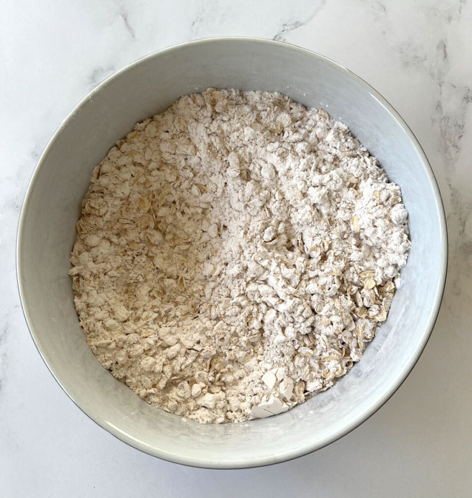 Large white mixing bowl with a dry white mixture of flour and oats in it. The bowl is on a white marble surface.