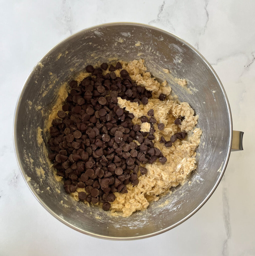 Stainless steel mixing bowl with oatmeal cookie dough in it with a pile of chocolate chips on top of the dough. Bowl is on a white marble surface.