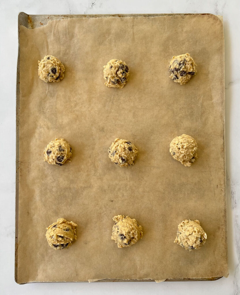 9 cookie dough balls on a parchment paper lined cookie sheet. Cookie sheet is on a white marble surface.