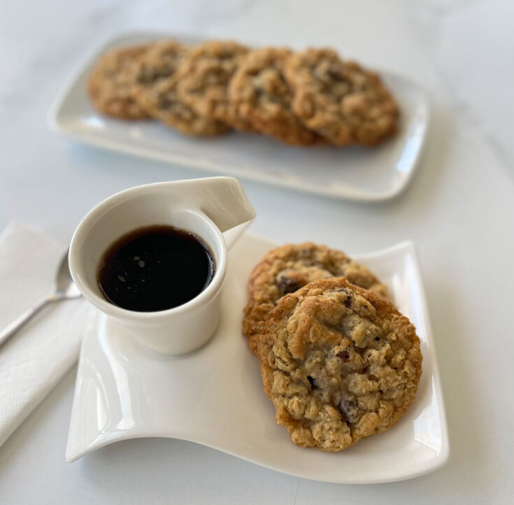 White rectangular plate with 2 oatmeal cookies and a small cup of coffee on it. There is a white napkin with a spoon resting on it on the left side of the plate. There is a white tray with 5 oatmeal cookies on it in the background. All the items are on a white marble surface.