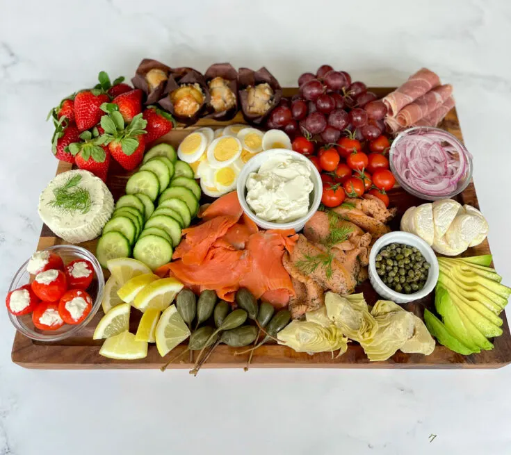 A large rectangle wood board with the following on it: sliced smoked salmon, sliced lox, sliced cucumber, sliced hard boiled eggs, sliced avocado, 6 slices of prosciutto rolled up, mini brie rounds cut in half, jarred artichoke hearts, caper berries, whole strawberries, cherry tomatoes, red seedless grapes, and lemon wedges. Also on the board there are 2 small clear round glass bowls. One contains thinly sliced red onion and the other contains small red round peppers stuffed with white cheese. There is a white round bowl with cream cheese and a smaller white round bowl with capers in it. There is a disc of herbed white cheese on the board. The board is on a white marble surface.