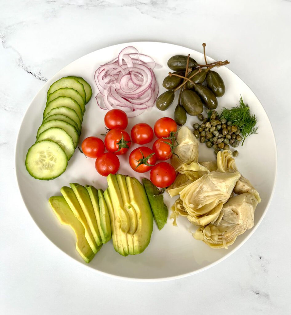 Large round white plate with the following on it: sliced cucumber, thinly sliced red onion, sliced avocado, 9 cheery tomatoes, 8 caper berries, a small pile of capers, chopped dill, and 6 jarred artichoke hearts. The large plate is on a white marble surface.