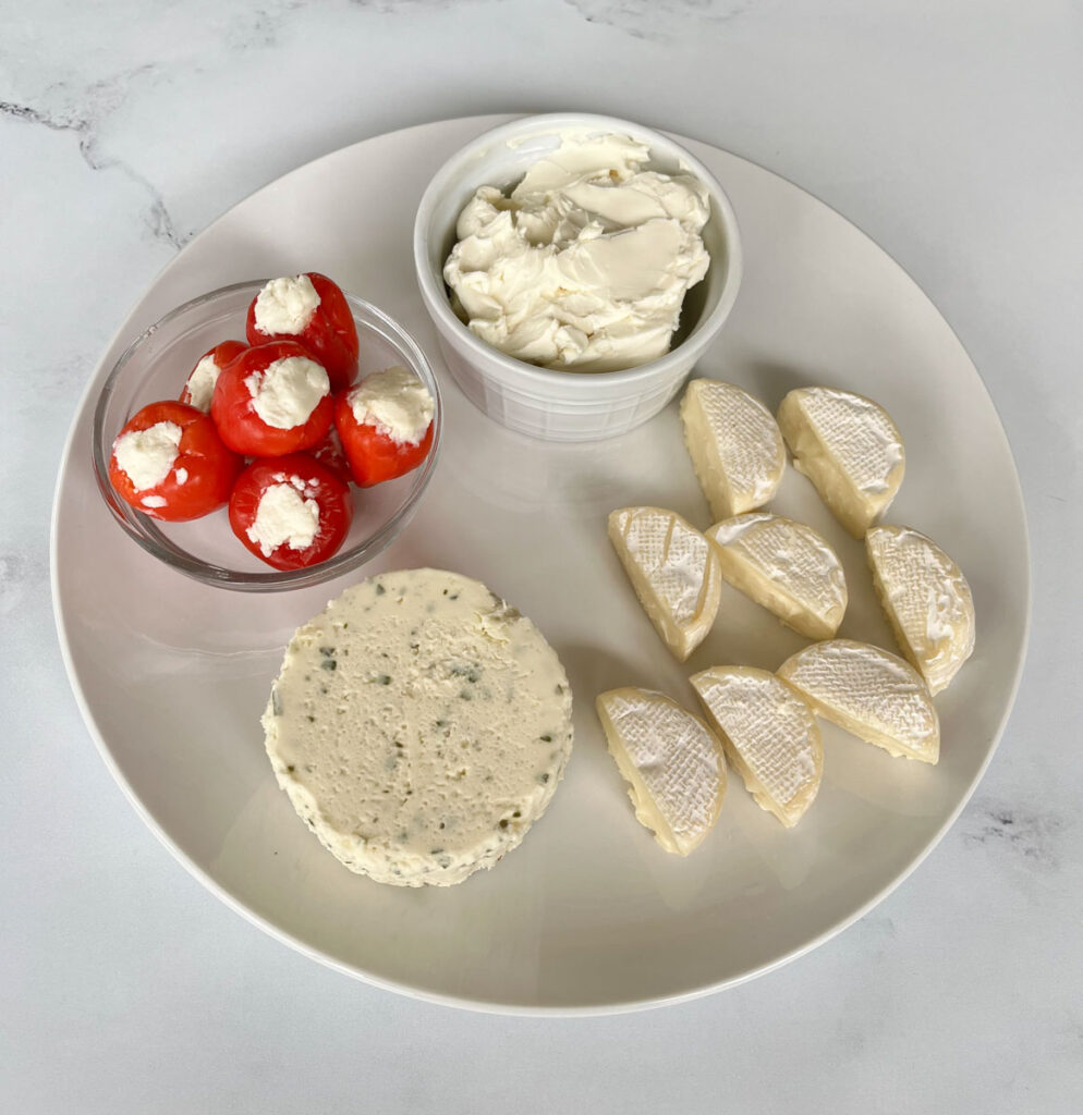 Large white round plate with a disc of white herbed cheese, eight half small discs of white brie cheese. There is a small white round bowl with cream cheese in it sitting on the plate. There is a small clear bowl with small red round peppers stuffed with cheese in it. The bowl is sitting on the plate. The large white plate is on a white marble surface.