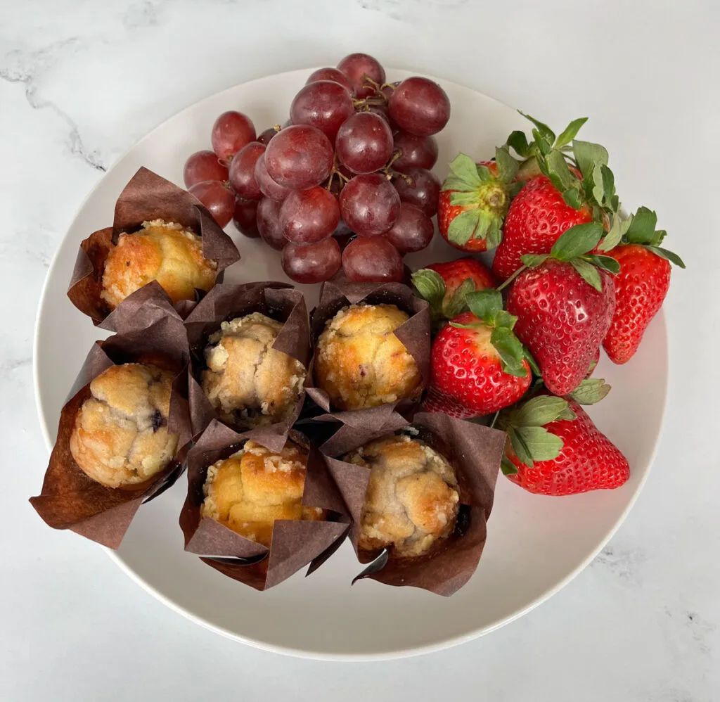 Large white round plate with whole strawberries, red grapes and blueberry mini muffins. Each muffin is set inside a brown paper cup. Plate is on a white marble surface.