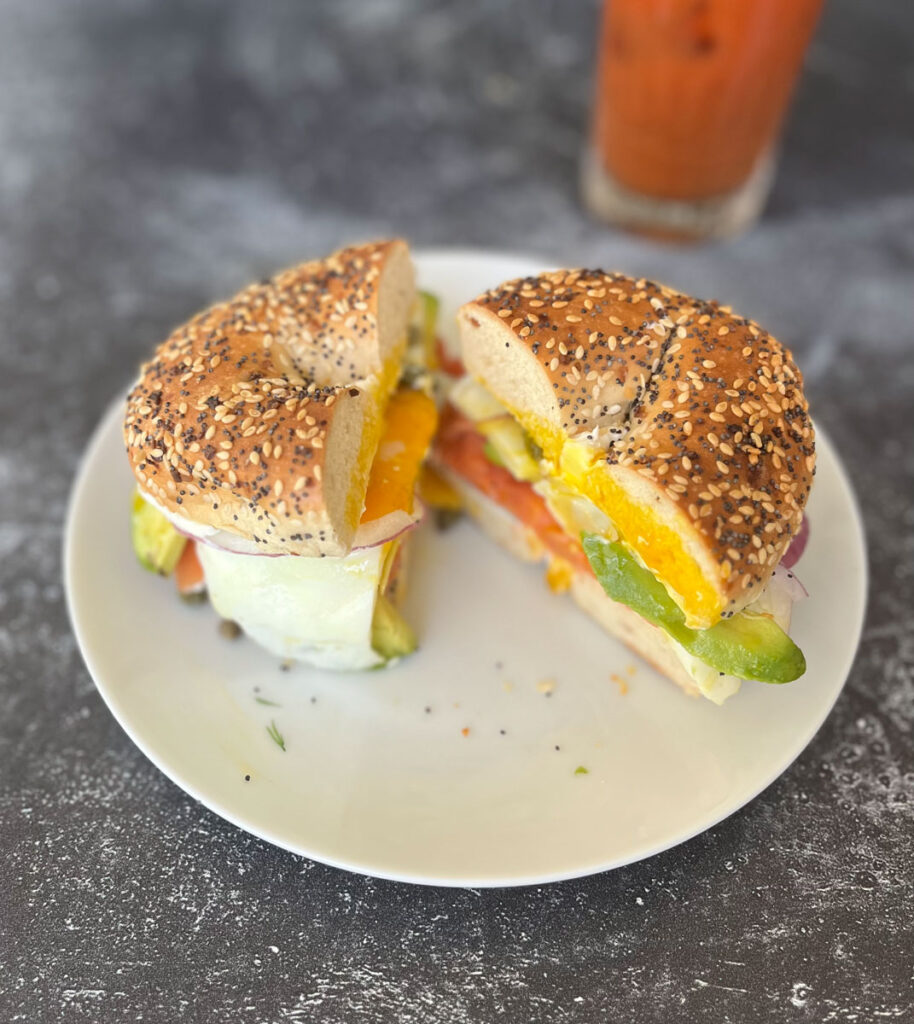 Bagel sandwich with lox, cream cheese, avocado, sliced cucumber, capers, and a fried egg. The sandwich is on a white round plate and cut in half. There is a clear tall glass with bloody mary mix in it. Items are on a gray surface.