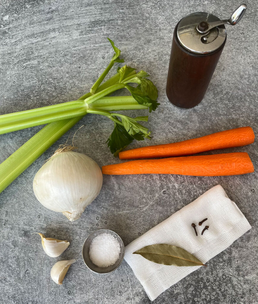 There are two stalks of celery, one white onion, 2 cloves of garlic, 2 peeled carrots, a small stainless steel ramekin with salt in it, a rectangular piece of white cheese cloth with a bay leaf and 3 cloves on it, and a wooden pepper grinder. All items are on a light gray surface.
