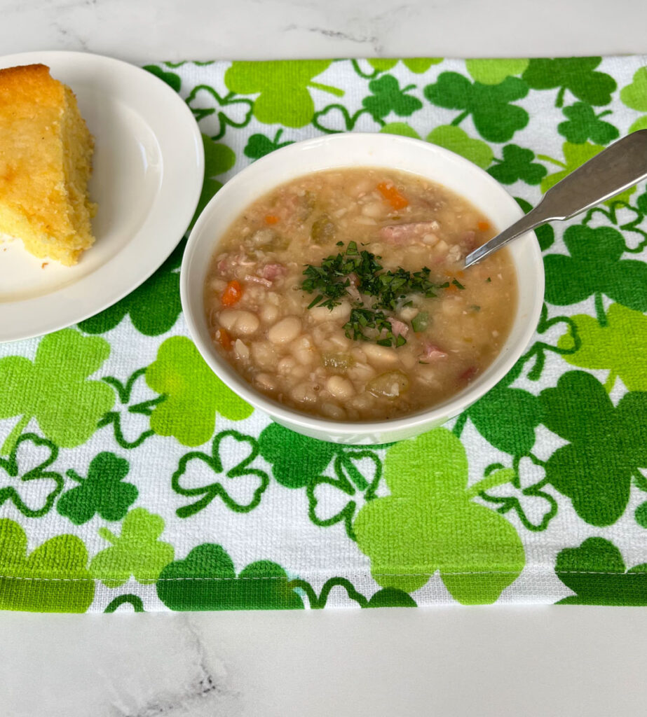Small white bowl with bean soup in it. There is chopped parsley on the soup with a spoon in it. There is a white round plate with a wedge of corn bread in the background. Dishes are on a white cloth with green shamrocks on it. The cloth is on a white marble surface.