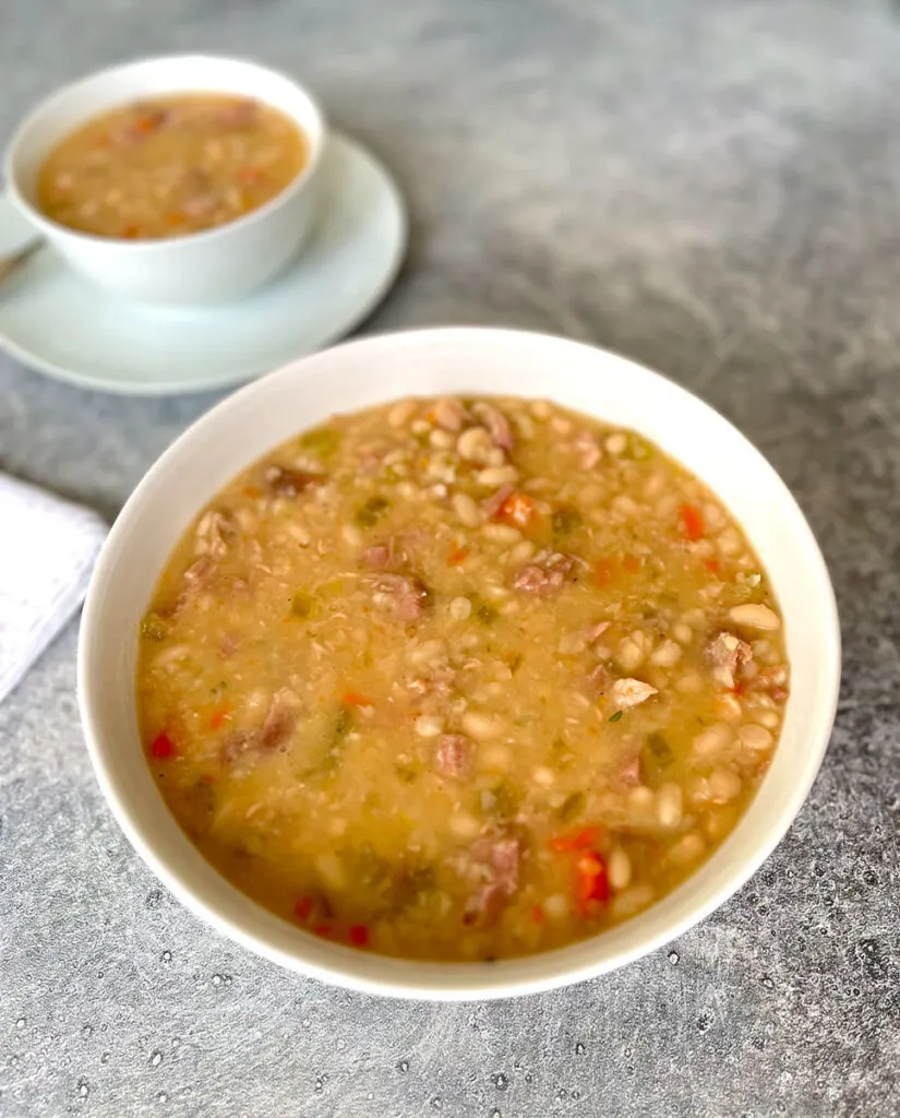Large white bowl of navy bean soup. There is a small white bowl sitting on a white round plate in the background. Items are on a light gray surface.