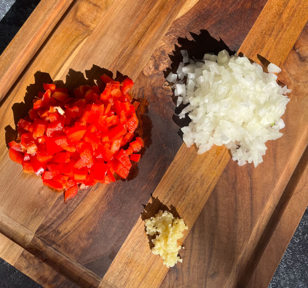 There is a wooden cutting board with a pile of chopped red pepper and a pile of chopped white onion. In front of the piles there is a small pile of minced garlic. The wooden cutting board is on a dark gray surface.