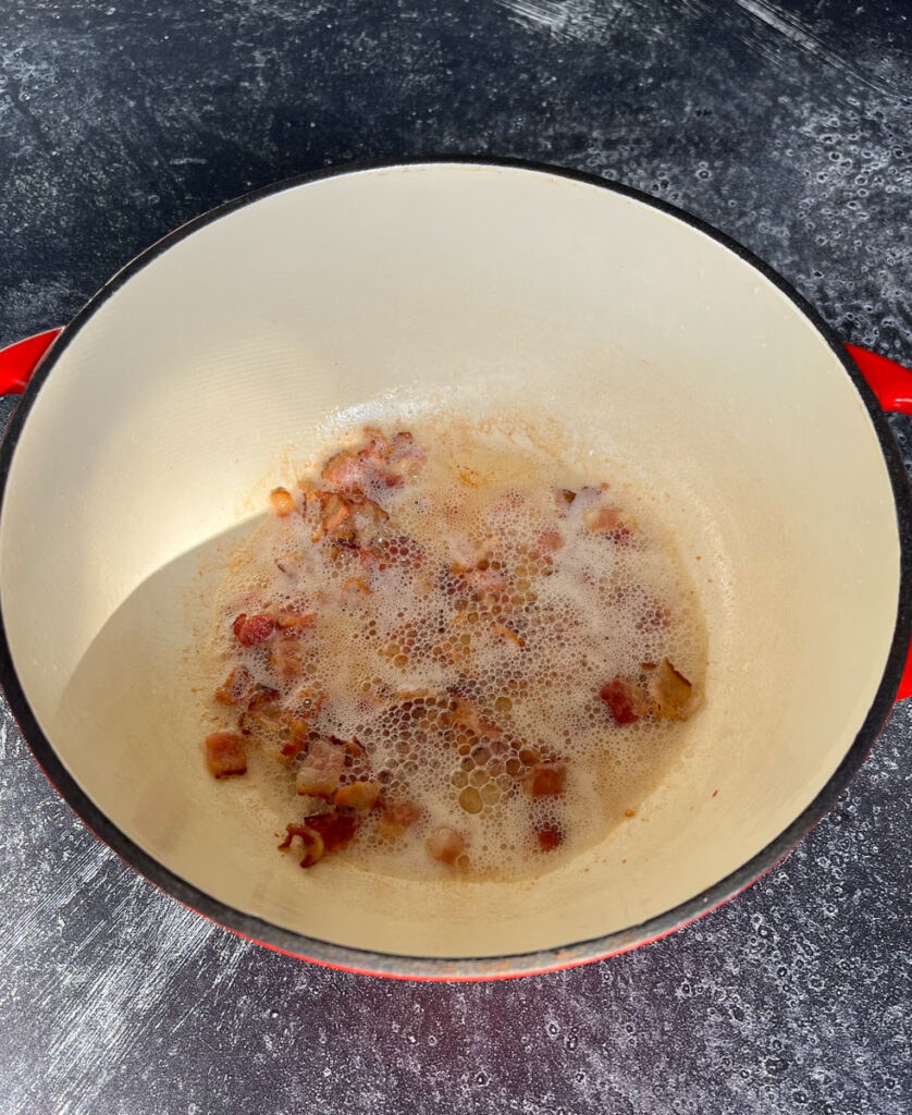 Large round pot with cut up bacon cooking in it. The pot is on a dark gray surface.