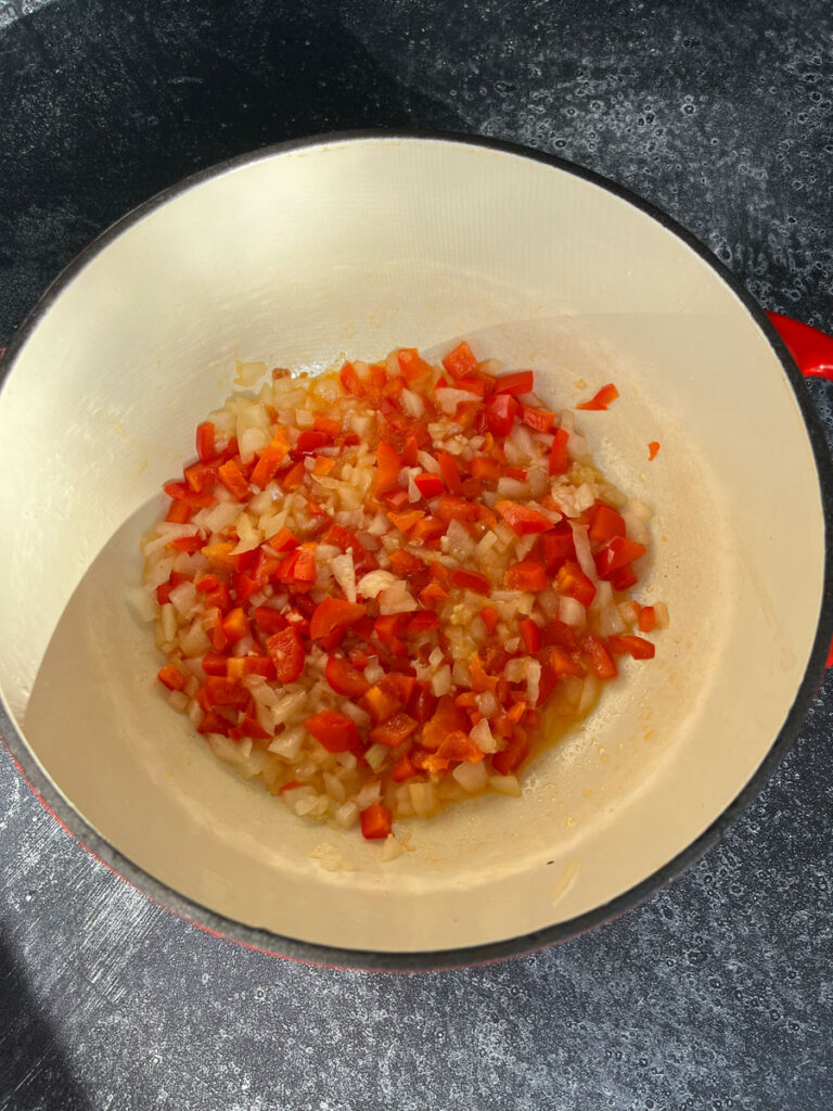 Large round pot with cut up red pepper, onion and garlic cooking in it. The pot is on a dark gray surface.