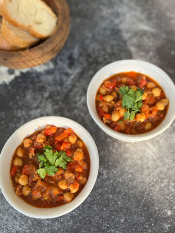 Two white round bowls with garbanzo beans, chorizo sausage, bacon and tomatoes in it. There is chopped fresh parsley on top of the beans. There is a wooden bowl with slices of white bread in the background. Bowls are on a dark gray surface.