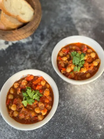 Two white round bowls with garbanzo beans, chorizo sausage, bacon and tomatoes in it. There is chopped fresh parsley on top of the beans. There is a wooden bowl with slices of white bread in the background. Bowls are on a dark gray surface.
