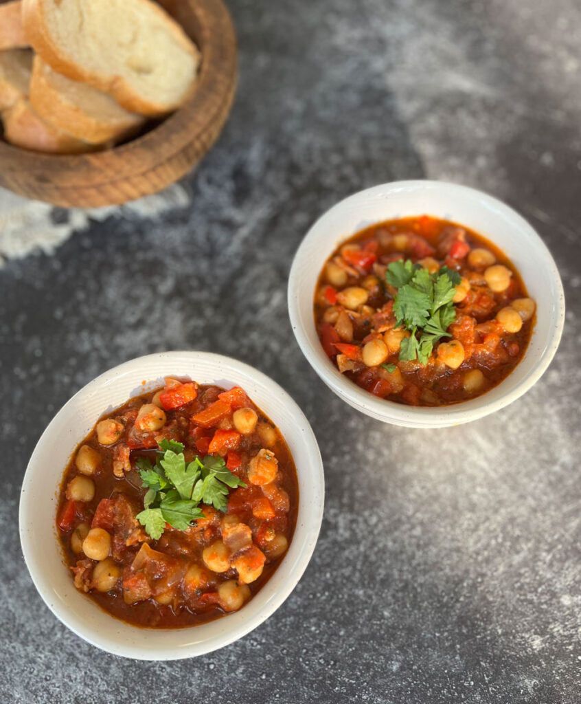 Two white round bowls with garbanzo beans, chorizo sausage, bacon and tomatoes in it. There is chopped fresh parsley on top of the beans. There is a wooden bowl with slices of white bread in the background. Bowls are on a dark gray surface.