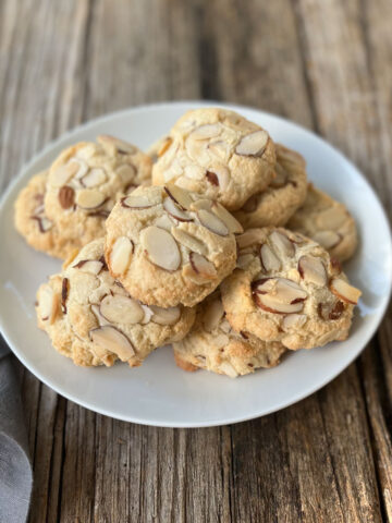 Round white plate with 9 beige cookies with sliced almonds on them. There is a gray cloth napkin next to the plate. The items are on a wood surface.
