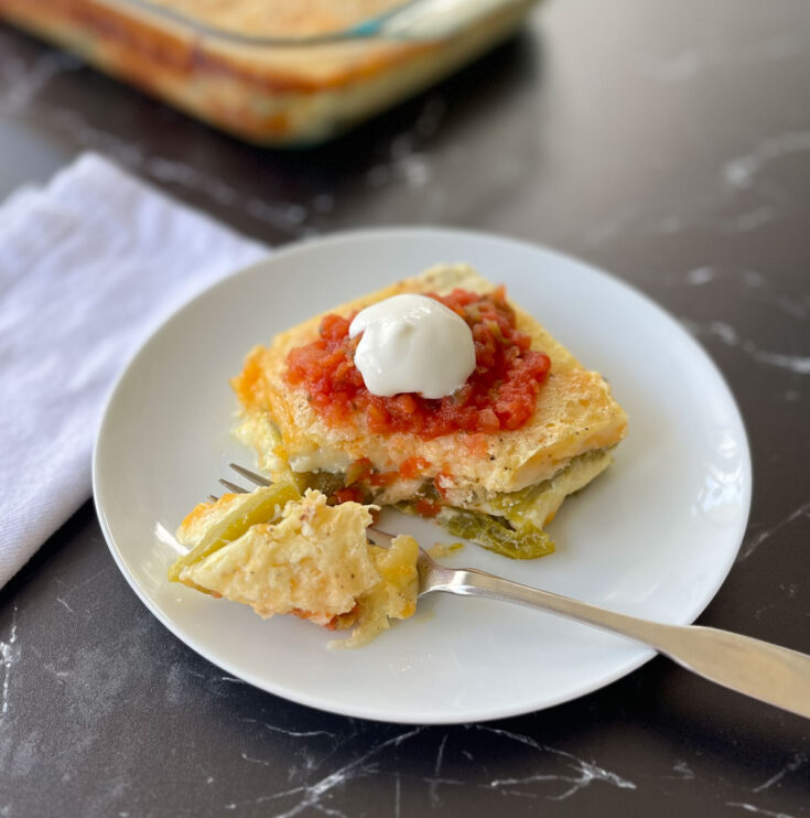 There is a white round plate with a piece of egg, cheese, and green chilis casserole on it. There is red salsa and a dollop of sour cream on top of the casserole. There is a fork on the plate with some of the casserole on it. There is a white cloth napkin on the side of the plate. In the background the is a clear baking dish with casserole in it. All the items are on a black marble surface.