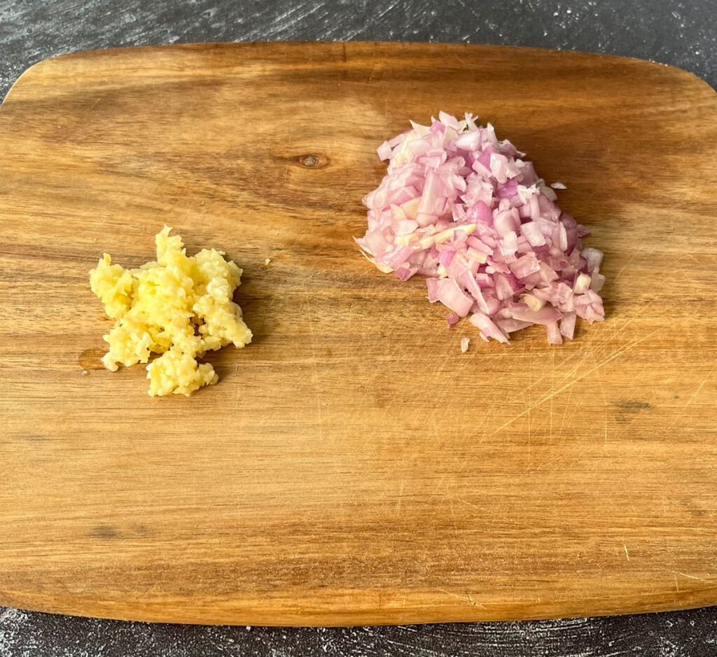 There is a small rectangular wooden cutting board with a pile of chopped shallots and a smaller pile of minced garlic on it. The cutting board is on a dark gray surface.