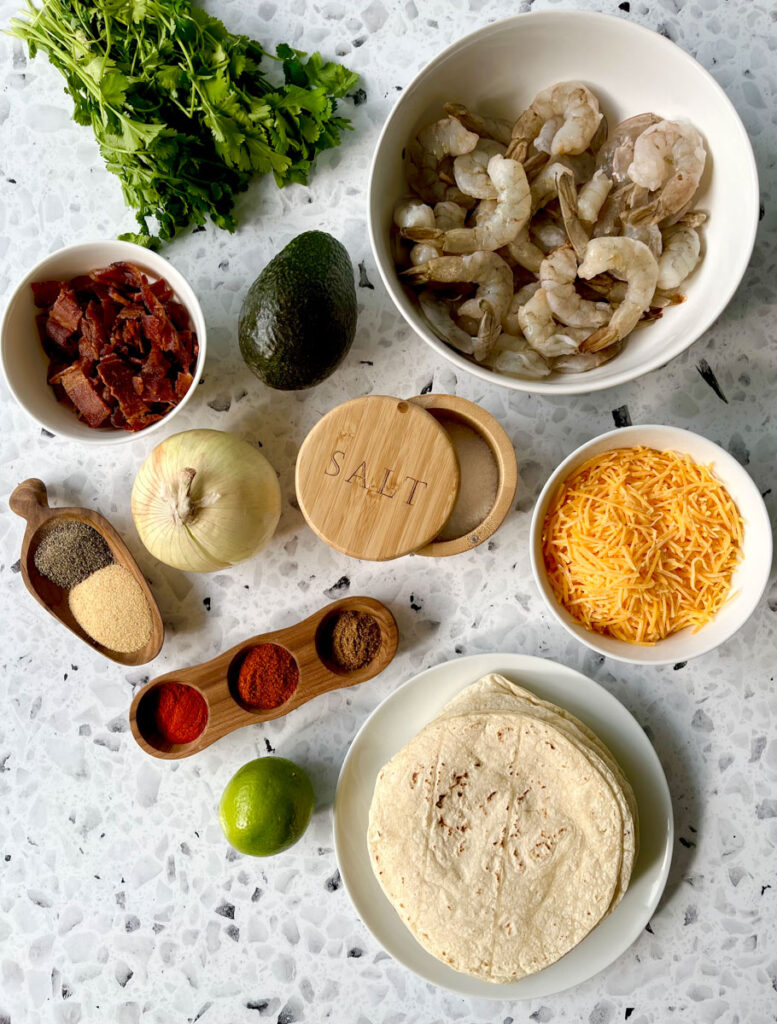 There is a white bowl with raw shrimp, a white bowl with shredded yellow cheese in it. a white bowl with cooked crumbled bacon in it. There is an avocado, onion, lime and a bundle of fresh cilantro. There is a white plate with corn tortillas on it. there is a wooden scoop with ground pepper and garlic powder in it. There is a wood vessel with three cups in it with red ground powder in the cups. There is a wood bowl with salt in it. All items are on a white and black terrazzo surface.
