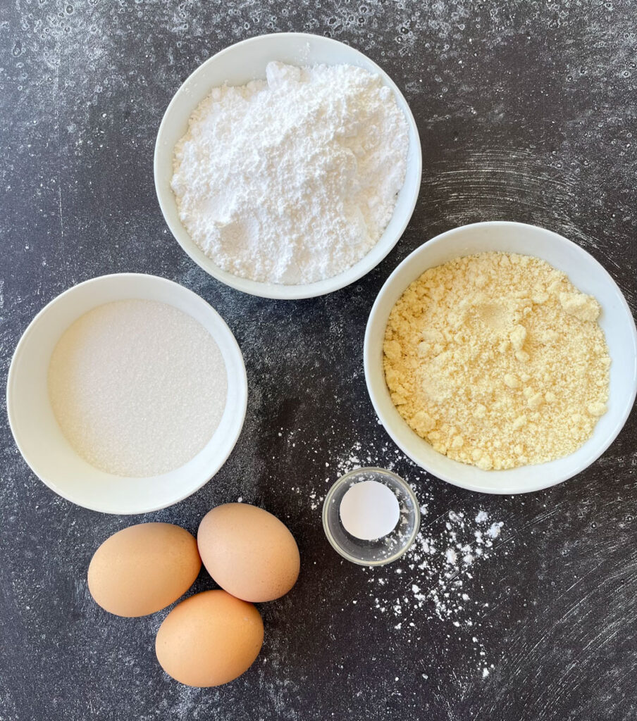 There are 3 white round bowls. One has white powdered sugar in it, one has white granulated sugar in it, and one has yellow almond flour in it. There are 3 brown eggs and a small clear ramekin with white food coloring in it. All items are on a dark gray surface.