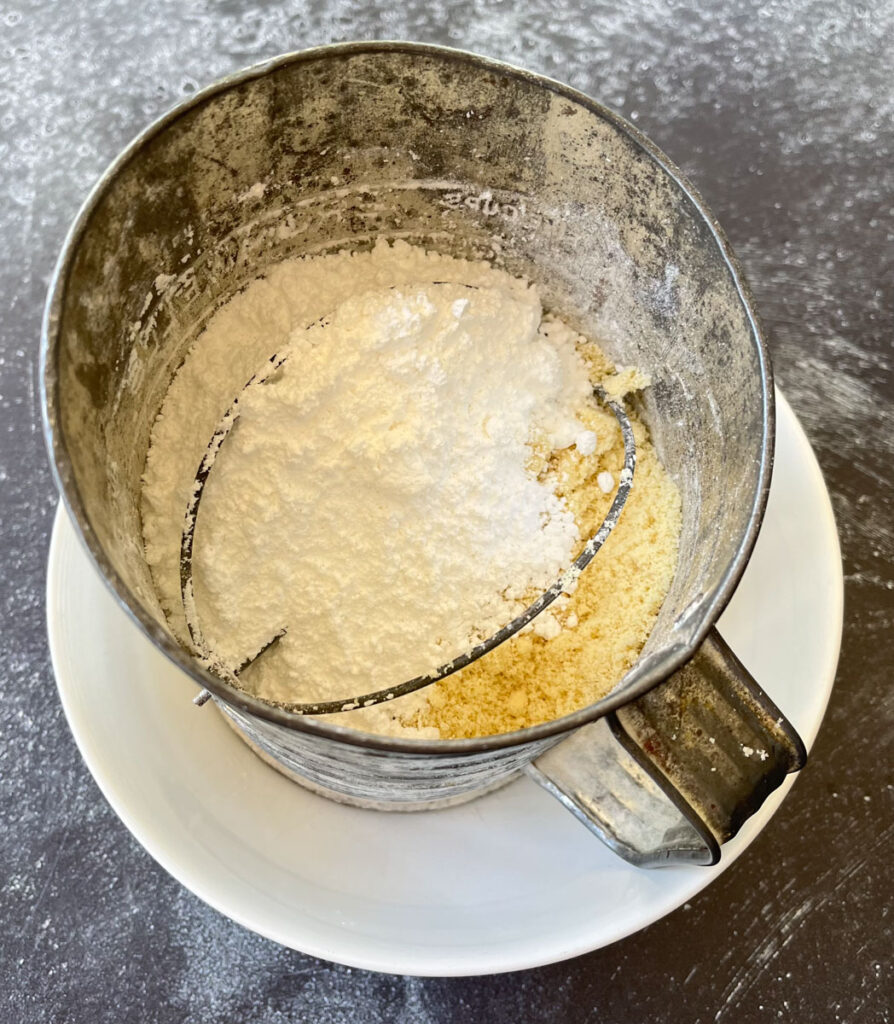 There is a stainless steel sifter sitting inside a large white bowl. There is white powdered sugar and yellow almond flour in the sifter. Bowl is sitting on a dark gray surface.