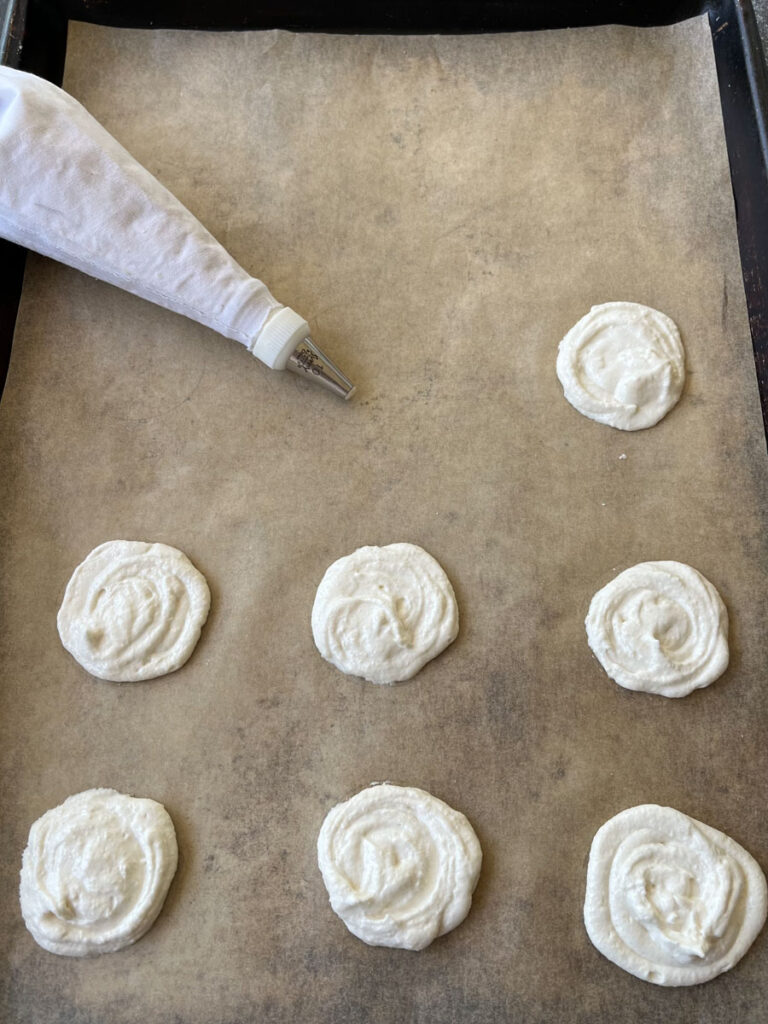 There is a baking pan lined with light brown parchment paper. There are seven round mounds of off white cookie batter on the parchment paper. There is a white piping bag with a silver tip on it sitting on the parchment paper in the background.