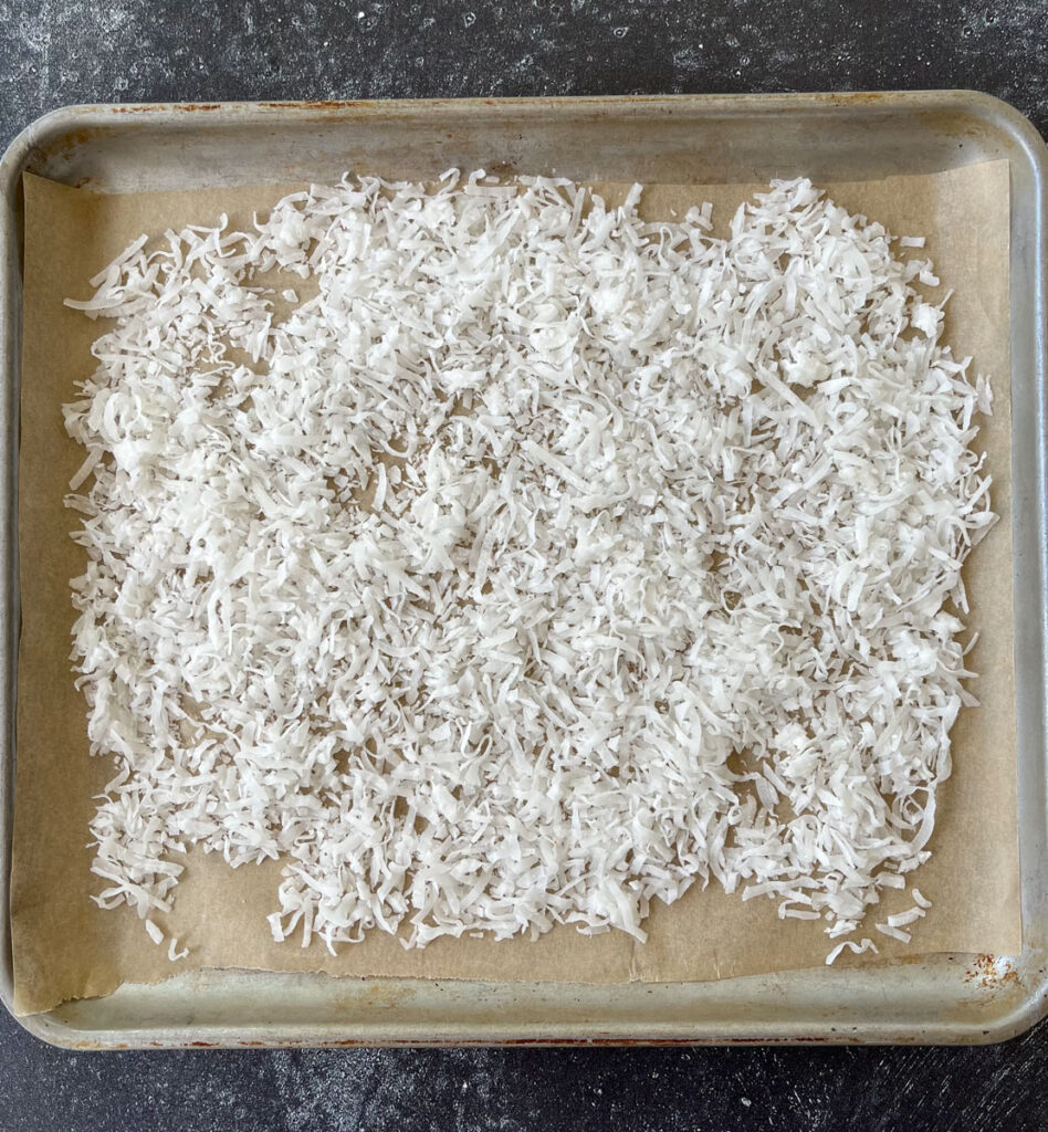 There is a baking pan lined with light brown parchment paper. There is white shredded coconut on the parchment paper. Baking pan is on a dark gray surface.