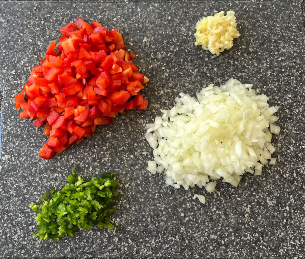 There is a mound of chopped up red bell pepper, a mound of chopped up onion, a smaller mound of finely chopped green jalapeno pepper, and a smaller mound of minced garlic. All items are on a speckled gray surface.