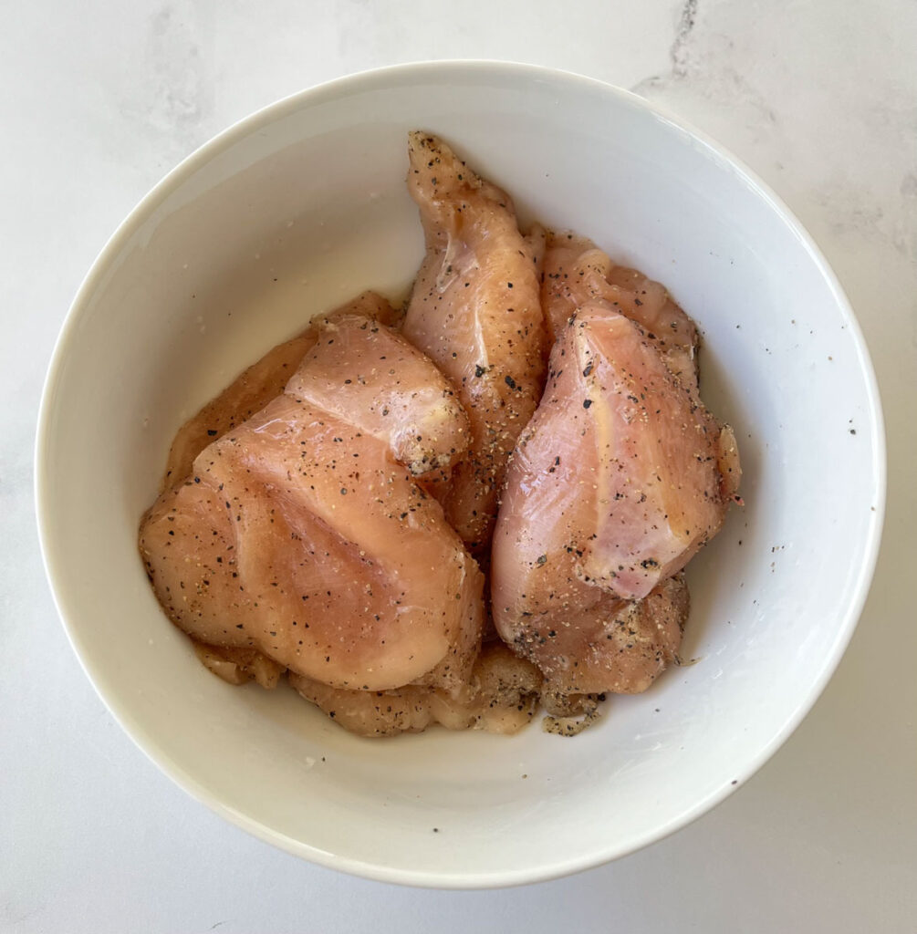 White large bowl with pieces of raw boneless skinless chicken breasts in it. The chicken has salt and pepper on it. The bowl is sitting on a white marble surface.