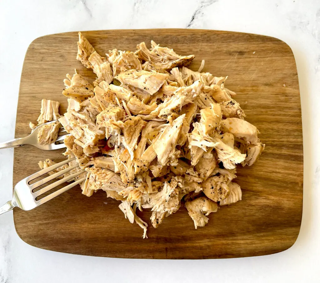 Cooked shredded chicken breast on a rectangular wood cutting board. There are 2 stainless steel forks leaning on the board. Board is on a white marble surface.