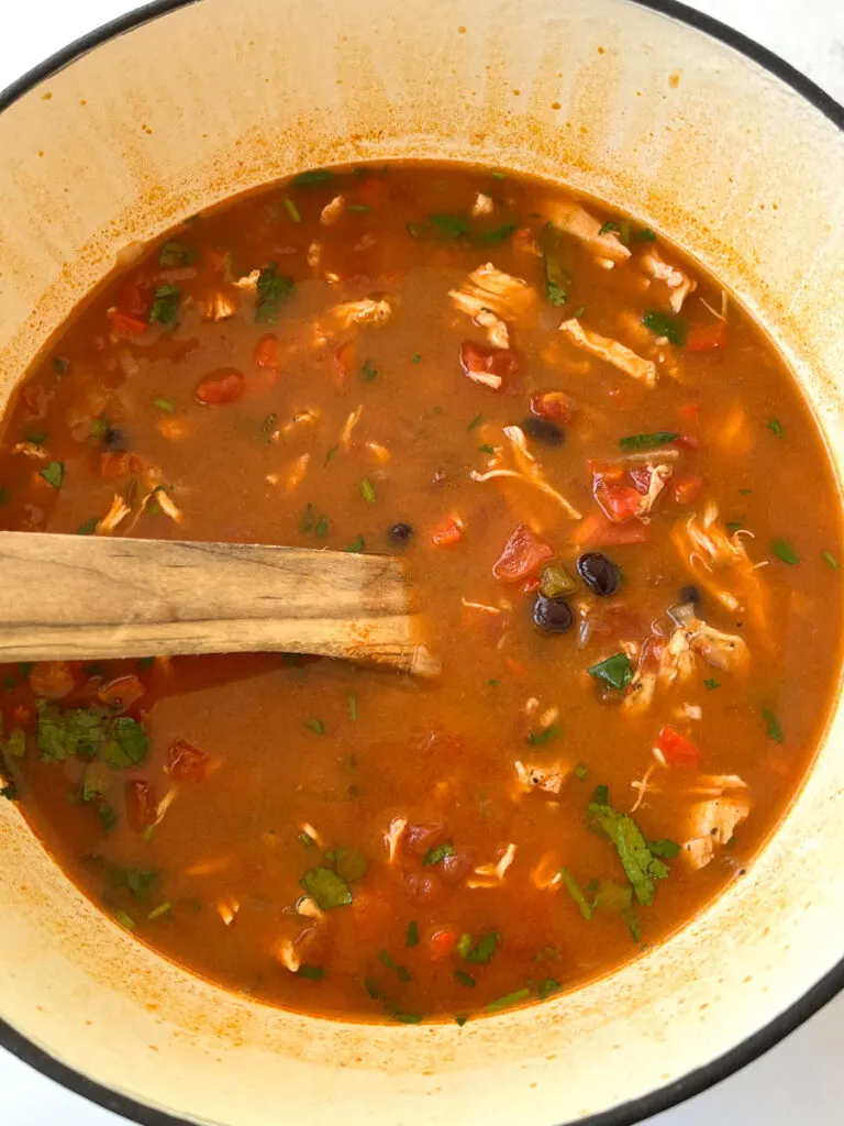 Large cream pot with shredded chicken, red sauce, chopped onion and peppers, black beans, chopped green cilantro, and chicken in it. There is a wooden spoon resting on the inside of the pot.