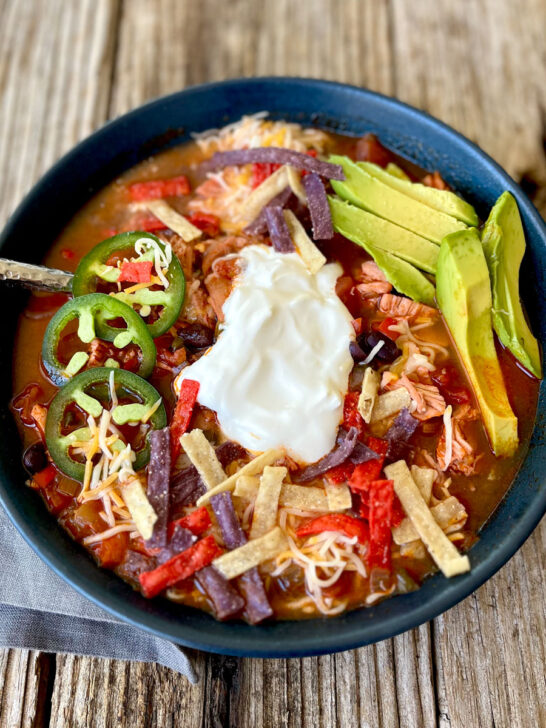 Blue bowl with red sauce, shredded chicken, sliced avocado, shredded cheese, a small mound of white sour cream in it. There is a stainless steel spoon in the bowl of soup. There is a gray cloth napkin laying next to the bowl. Items are on a wood surface.