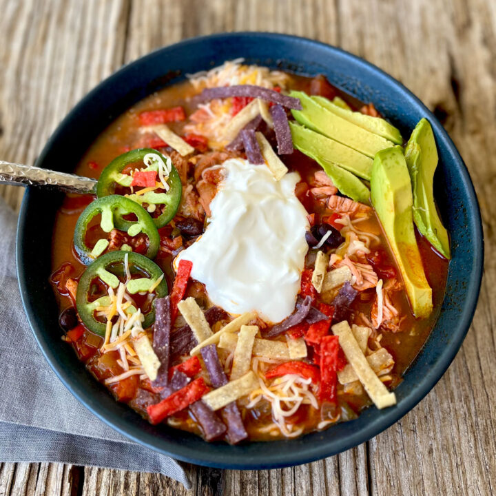 Blue bowl with red sauce, shredded chicken, sliced avocado, shredded cheese, a small mound of white sour cream in it. There is a stainless steel spoon in the bowl of soup. There is a gray cloth napkin laying next to the bowl. Items are on a wood surface.