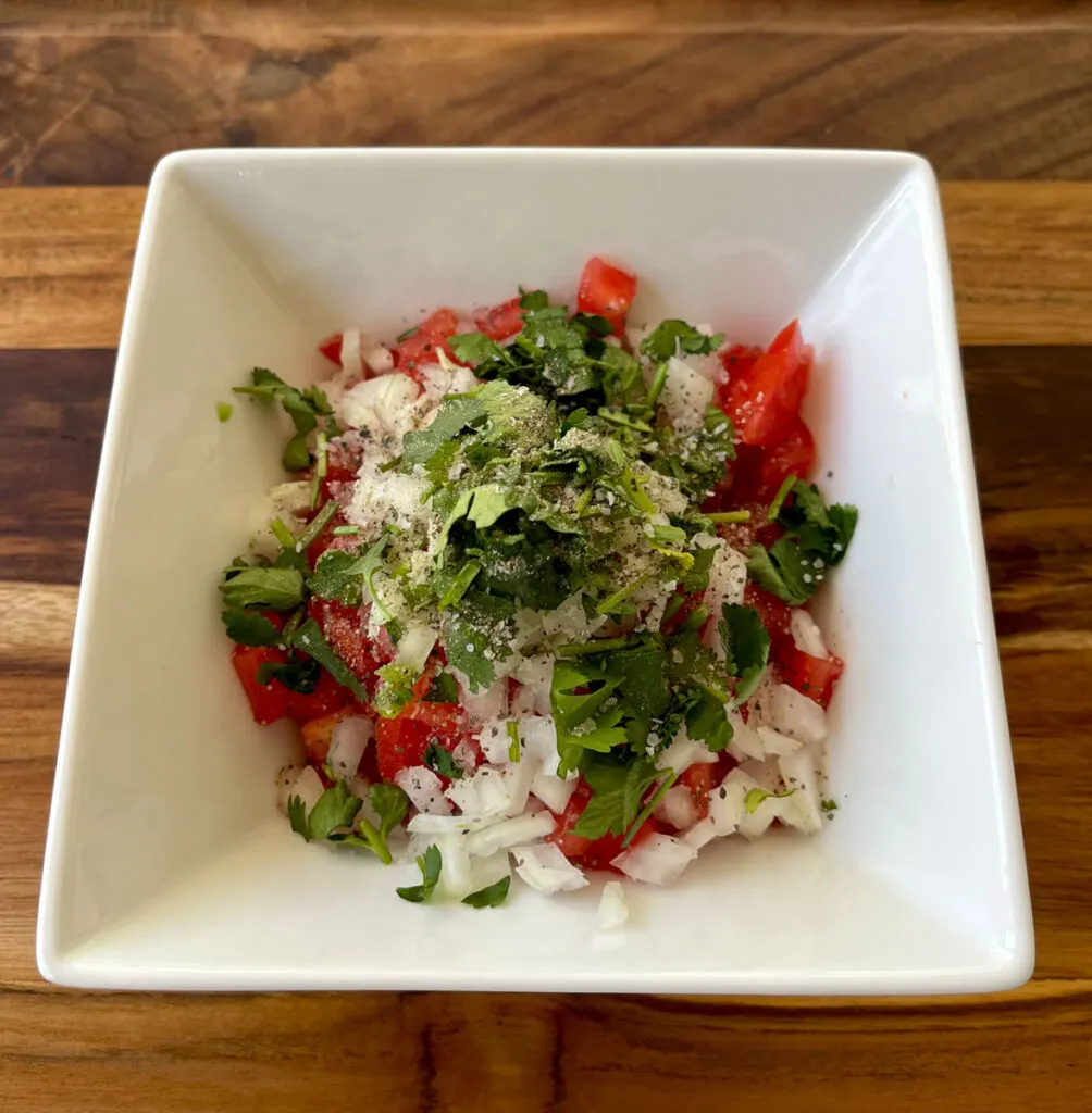 There is a square white bowl with chopped tomato, onion, jalapeno pepper, and green cilantro in it. The bowl is on a wood surface.
