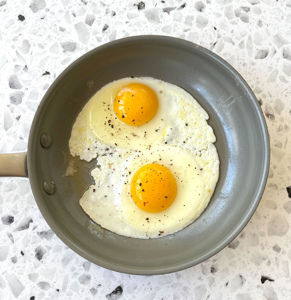 There is a gray fry pan with 2 sunny side up eggs with ground pepper on them. The pan is on a white and gray terrazzo surface.