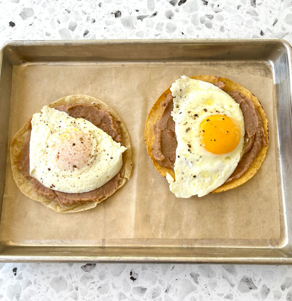 There is a stainless steel tray lined with brown paper. There is a white corn tortilla with refried beans and a fried egg with ground pepper on it. There is a yellow corn tortilla next to the white corn tortilla. There is refried beans and a sunny side up egg with ground pepper on it. The tray is on a white and black terrazzo surface.