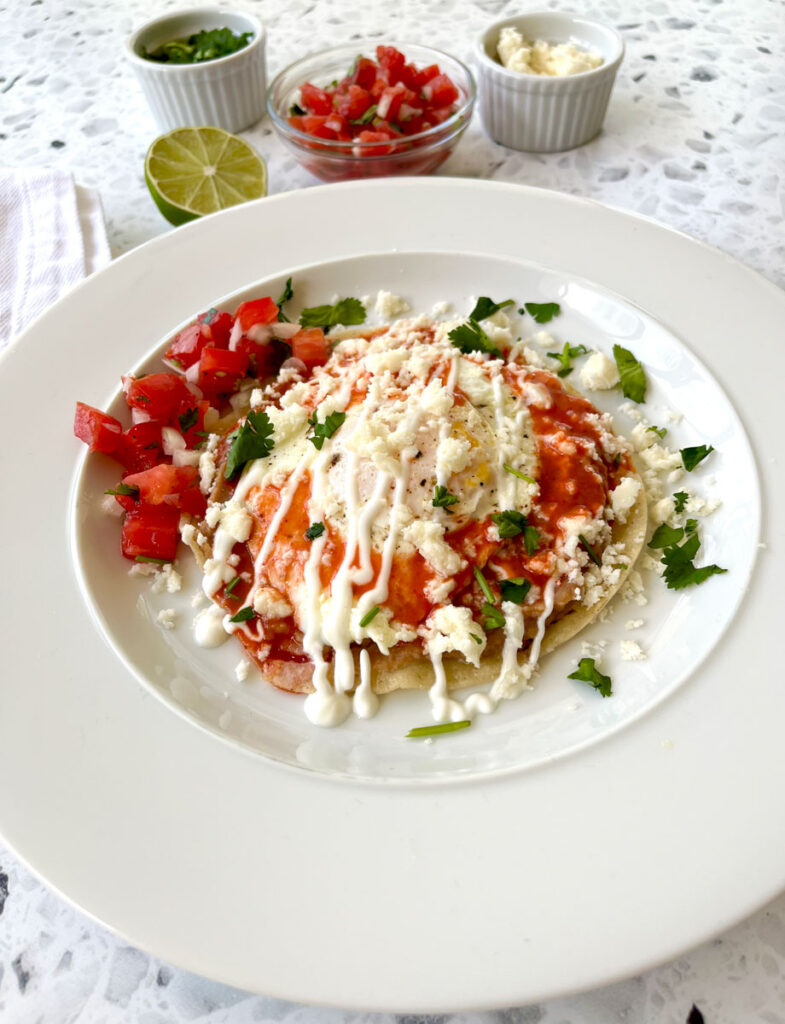 There is a white round plate with a white corn tortilla on it. There is refried beans, red sauce, a fried egg, crumbled white cheese, drizzled sour cream, ground pepper, and chopped green cilantro on the corn tortilla. There is a small clear bowl with chopped up tomato and onion in it, a small white bowl with crumbled white cheese in it, a second small white bowl with chopped green cilantro, and a half of a lime in the background. There is a white cloth napkin next to the plate. All items are in a white and black terrazzo surface.