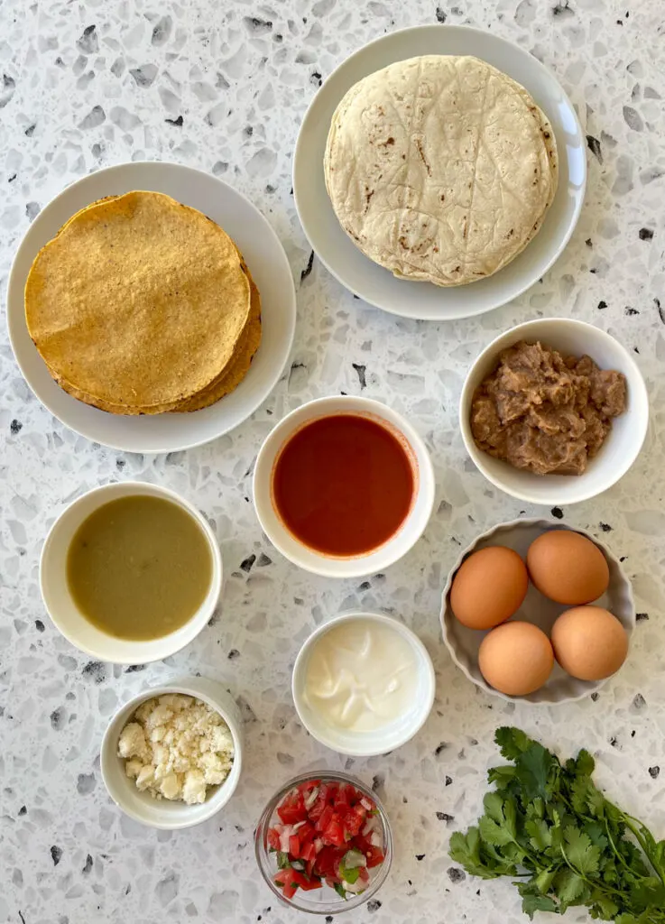 There are 2 white round plates. One has yellow hard corn tortillas on it, the other has white soft corn tortillas on it. There are 6 small bowls, one with green sauce, one with red sauce, one with light brown refried beans in it, one with four brown eggs in it, one with white crumbled cheese in it, and one with sour cream in it. There is a small clear bowl with chopped tomato and onion in it. There is a bunch of cilantro in the foreground. All items are on a white and black terrazzo surface.