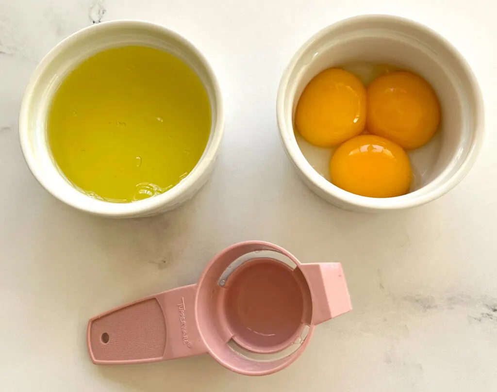 There are 2 small white bowls. One bowl has egg whites in it, the other bowl has 3 egg yolks in it. There is a pink plastic egg separator lying in front of the bowls. Items are on a white marble surface.