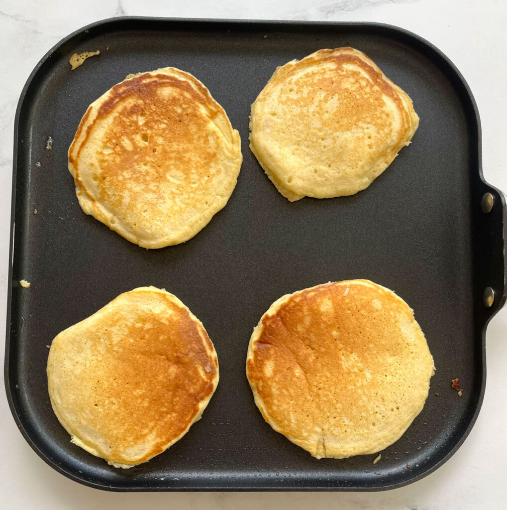 There are 4 golden brown pancakes on a rimmed square skillet. The skillet is on a white marble surface.