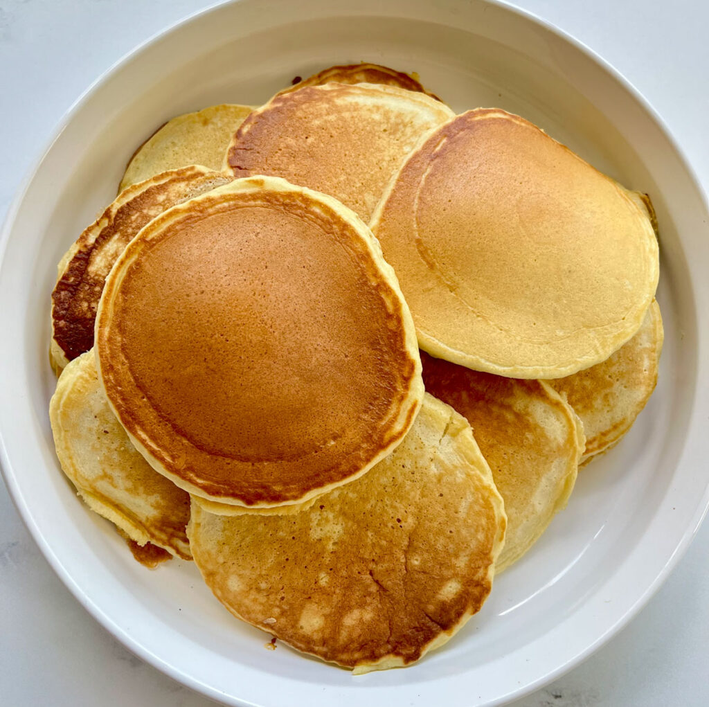 There is a large white serving dish with a mound of pancakes. The dish is on a white marble surface.