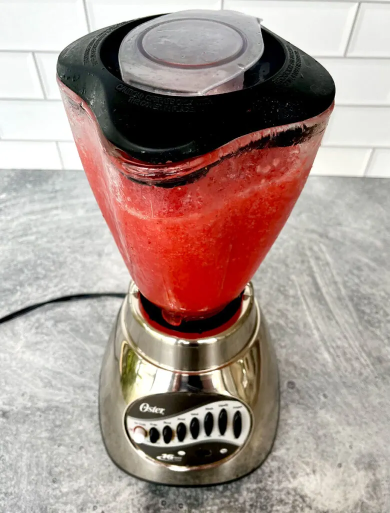 Stainless steel blender bass with a glass pitcher with a black lid on top of it. There is a red frozen slushy drink in the pitcher. The blender is sitting on a light gray surface.