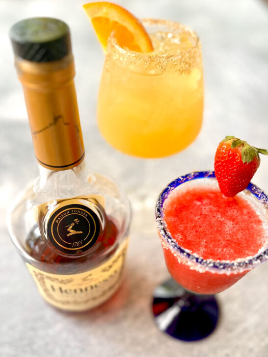 There is a stemmed clear glass with a blue rim. There is frozen strawberry margarita mix in the glass. The glass has sugar around the rim and a fresh strawberry on it. There is a second clear stemmed glass with light brown liquid and ice cubes in it. The rim of the glass has salt on it and an orange slice on it. There is a bottle of Hennessy Cognac next to the glasses. Items are on a light gray surface.