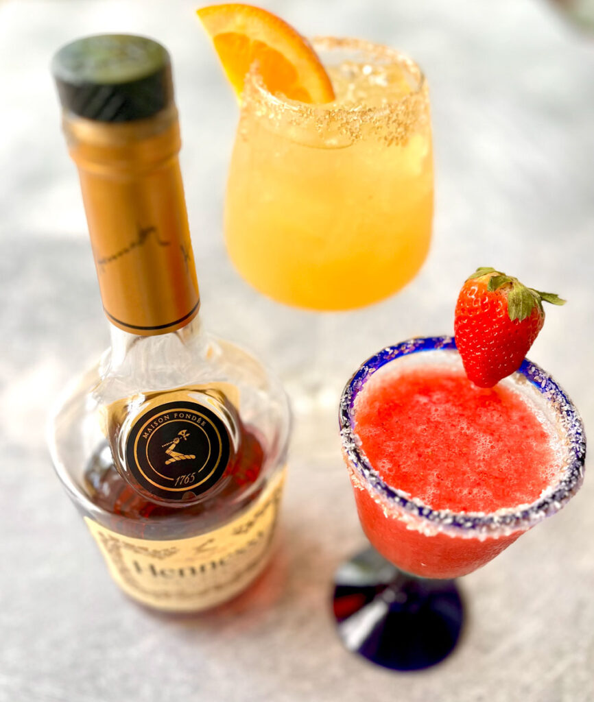 There is a stemmed clear glass with a blue rim. There is frozen strawberry margarita mix in the glass. The glass has sugar around the rim and a fresh strawberry on it. There is a second clear stemmed glass with light brown liquid and ice cubes in it. The rim of the glass has salt on it and an orange slice on it. There is a bottle of Hennessy Cognac next to the glasses. Items are on a light gray surface.